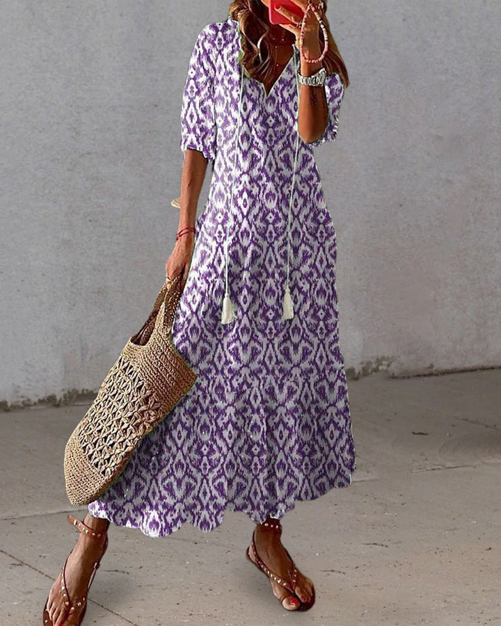 Woman wearing a purple patterned dress holding a woven bag against a plain background