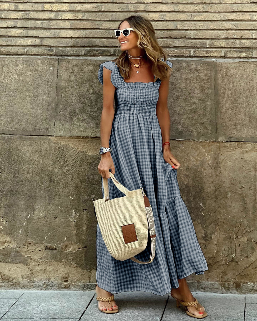 Woman in a blue checkered dress holding a straw bag against a stone wall.