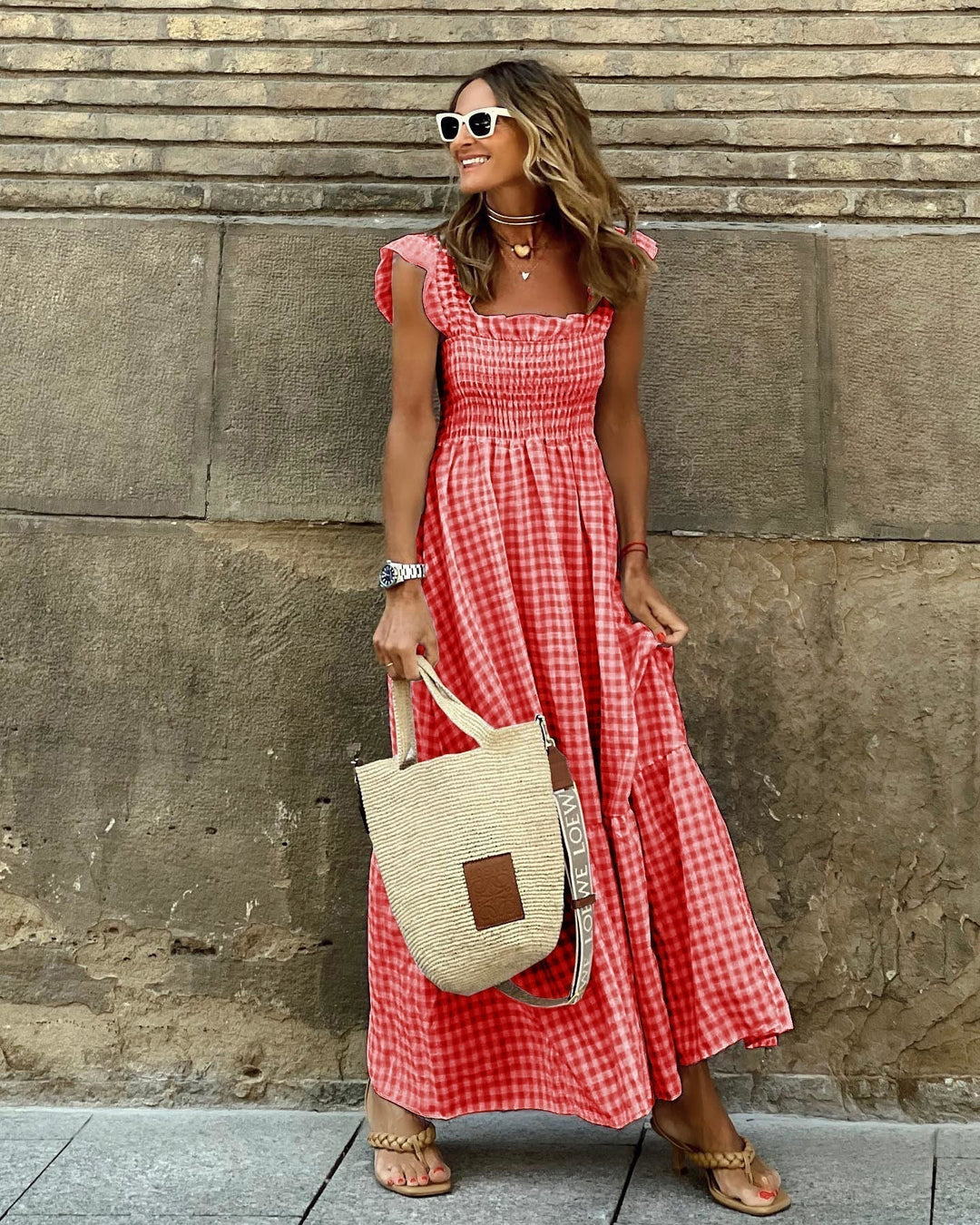 Woman in a red checkered dress holding a straw bag against a stone wall.