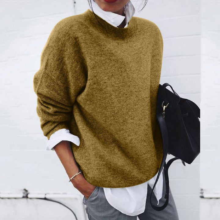 Person wearing a mustard yellow sweater with a white shirt and gray pants, holding a black backpack against a white background.