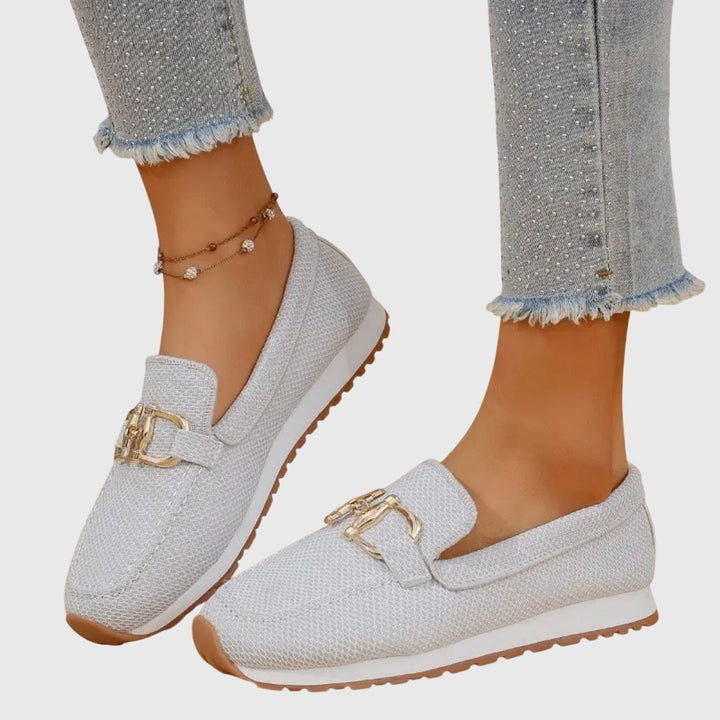 White loafers with gold buckles worn by a person on a light gray background