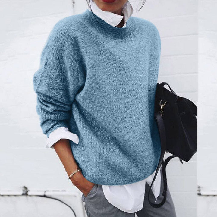 Person wearing a blue sweater with a white shirt and gray pants, holding a black backpack against a white background.