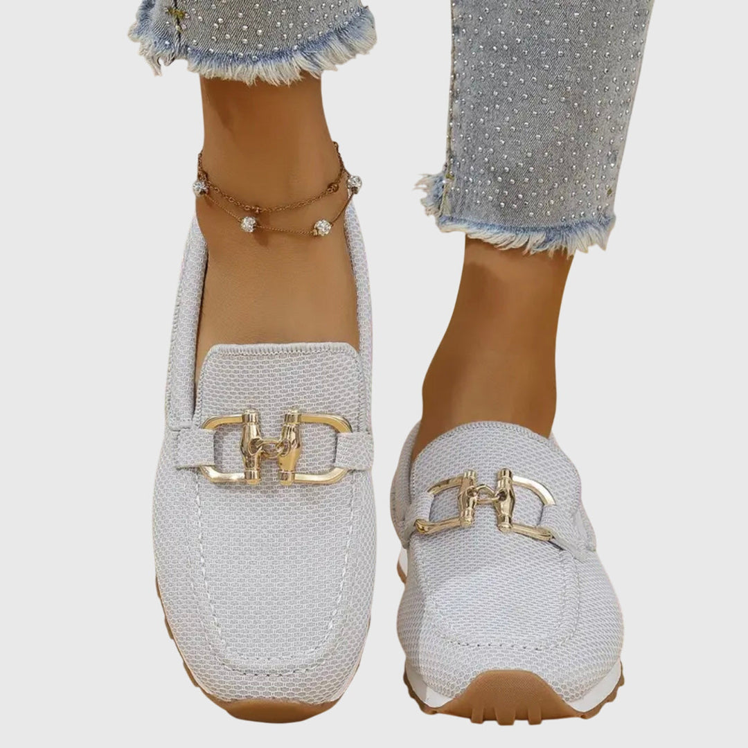 Light gray loafers with gold buckles worn by a person on a white background