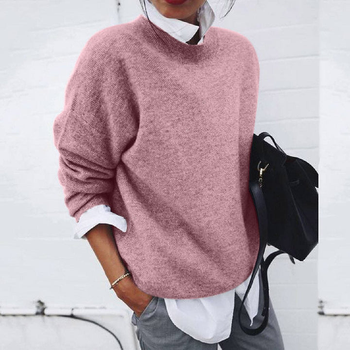 Person wearing a pink sweater with a white shirt and gray pants, holding a black bag against a white background.