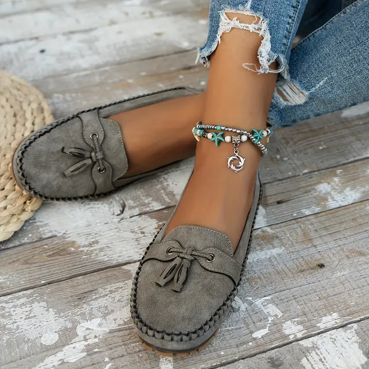 Gray loafers worn on a wooden floor with a person's leg and denim pants visible.