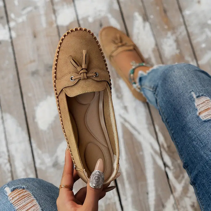 Brown suede loafer with a knot held by a hand on a wooden floor.