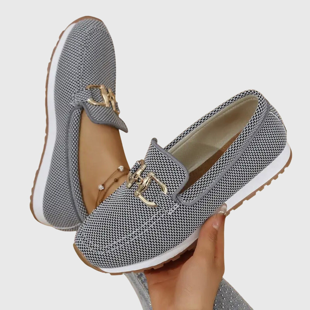 Gray patterned loafers with gold accents held by a hand on a light gray background