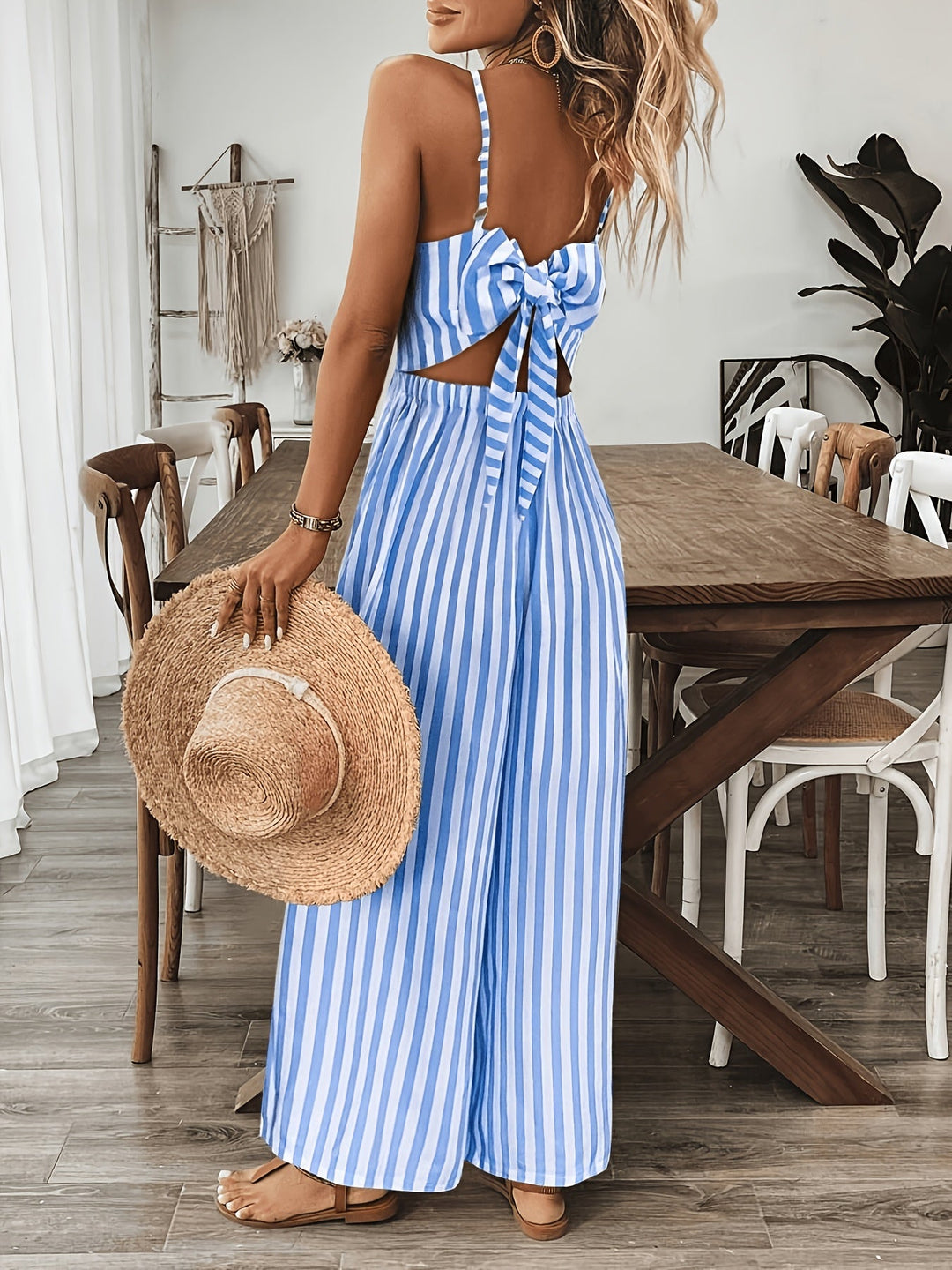Woman in a blue and white striped dress holding a straw hat in a room with wooden furniture.