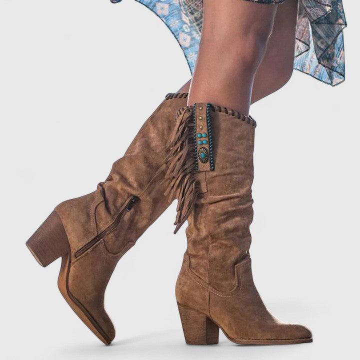 Brown suede boots with fringes and turquoise accents worn by a person on a white background