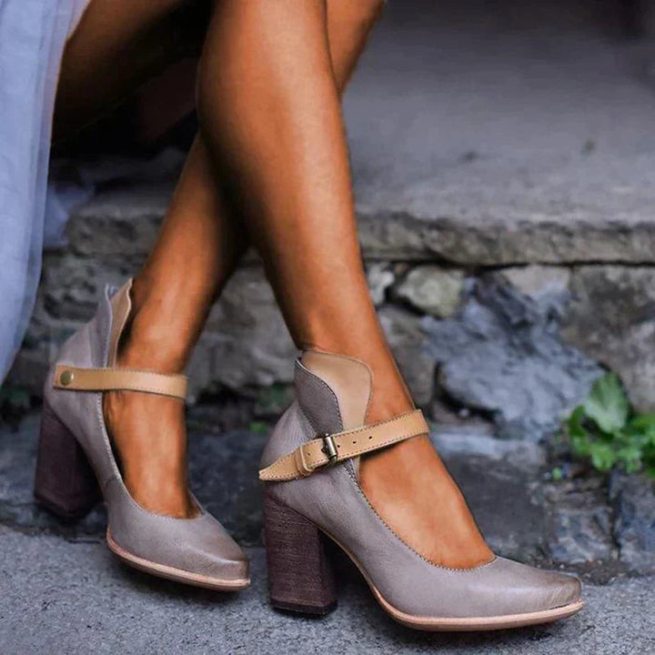 Gray high-heeled shoes with brown block heels worn by a person sitting on stone steps.