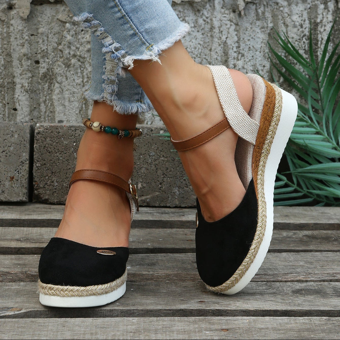 Black wedge sandals with brown straps worn by a person on a wooden floor.