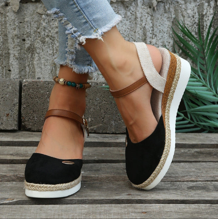 Black wedge sandals with brown straps worn by a person on a wooden floor.