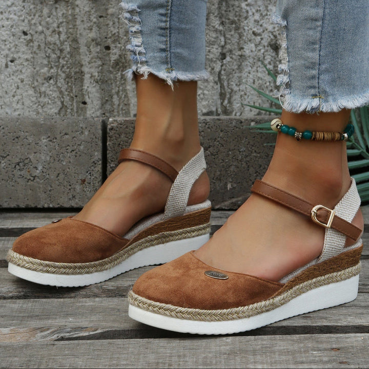 Brown suede sandals with white soles worn by a person on a wooden surface.