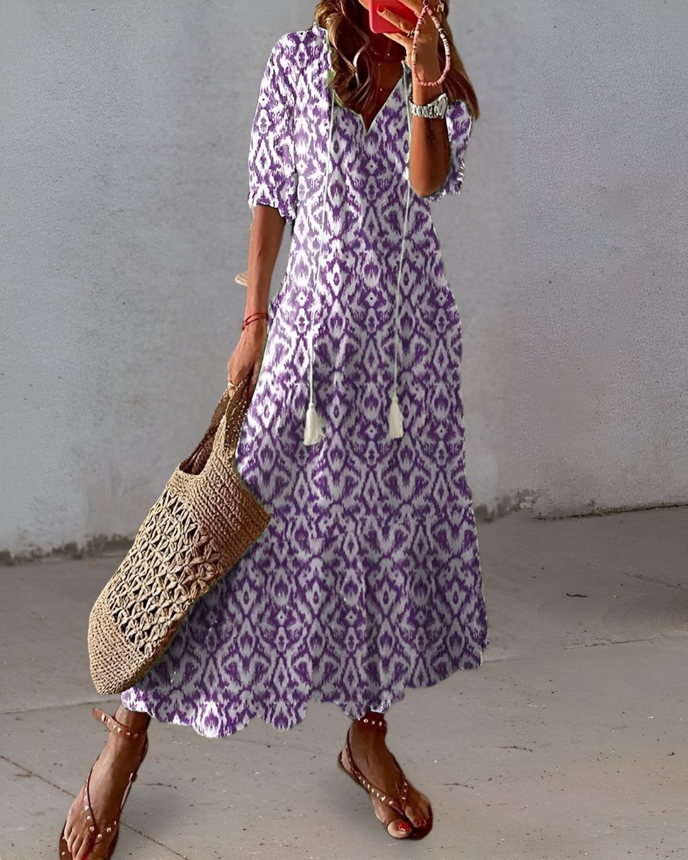 Woman wearing a long purple patterned dress holding a woven bag against a plain background