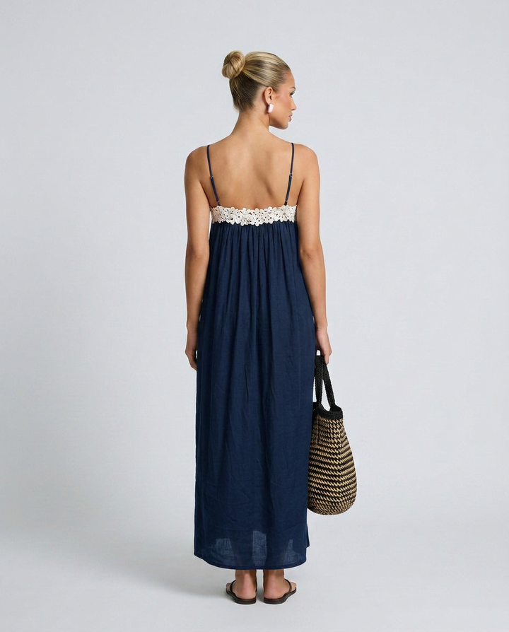 Woman wearing a navy blue dress with white lace trim, holding a straw bag on a light gray background