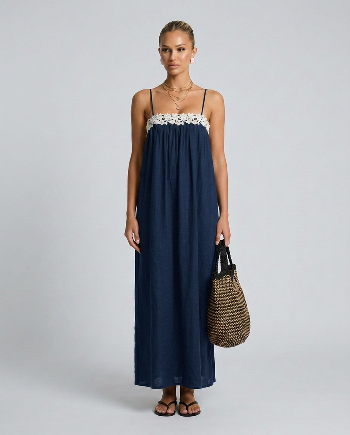 Woman wearing a navy blue dress with white floral details, holding a woven bag on a plain background