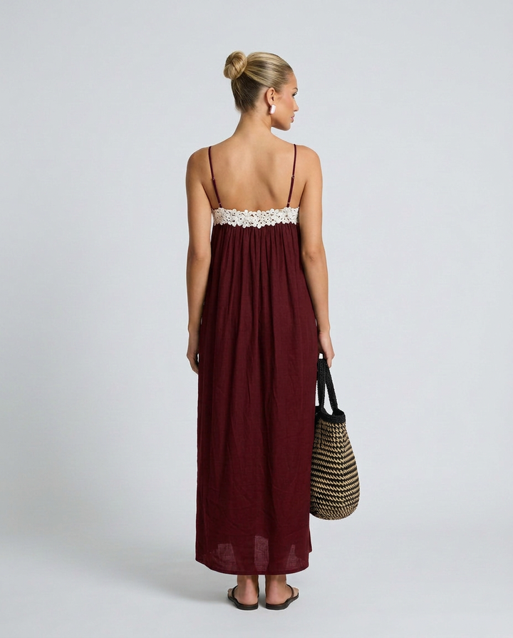 Woman wearing a burgundy dress with a white lace top, holding a straw bag on a plain background