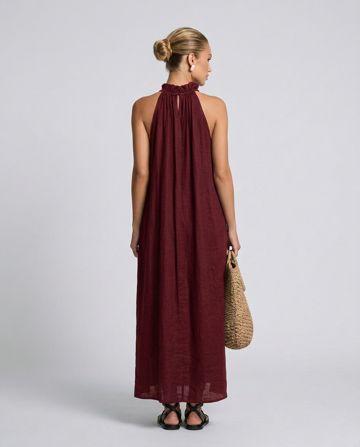 Woman wearing a burgundy halter neck dress holding a woven bag on a plain background