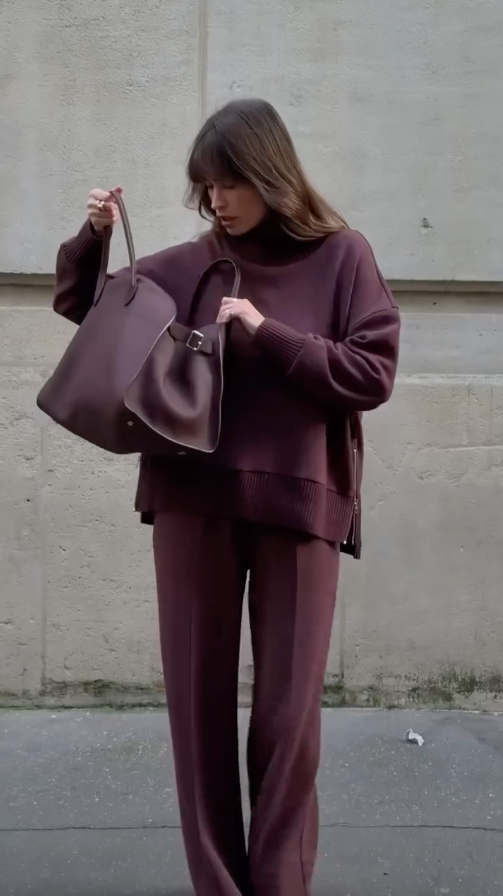 Person wearing a brown outfit holding a matching handbag against a plain background