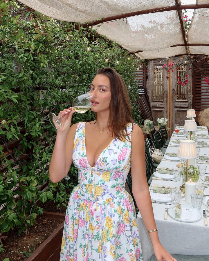 Woman in a floral dress holding a glass of white wine outdoors near a decorated table.