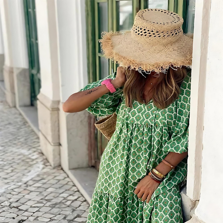 Woman in a green patterned dress and straw hat standing in front of a white wall.