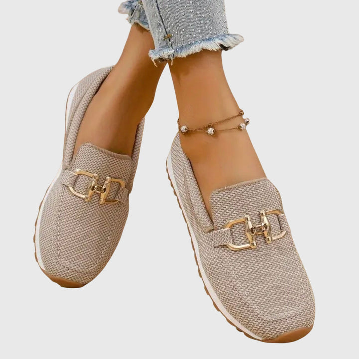 Beige loafers with gold buckles worn by a person on a light gray background