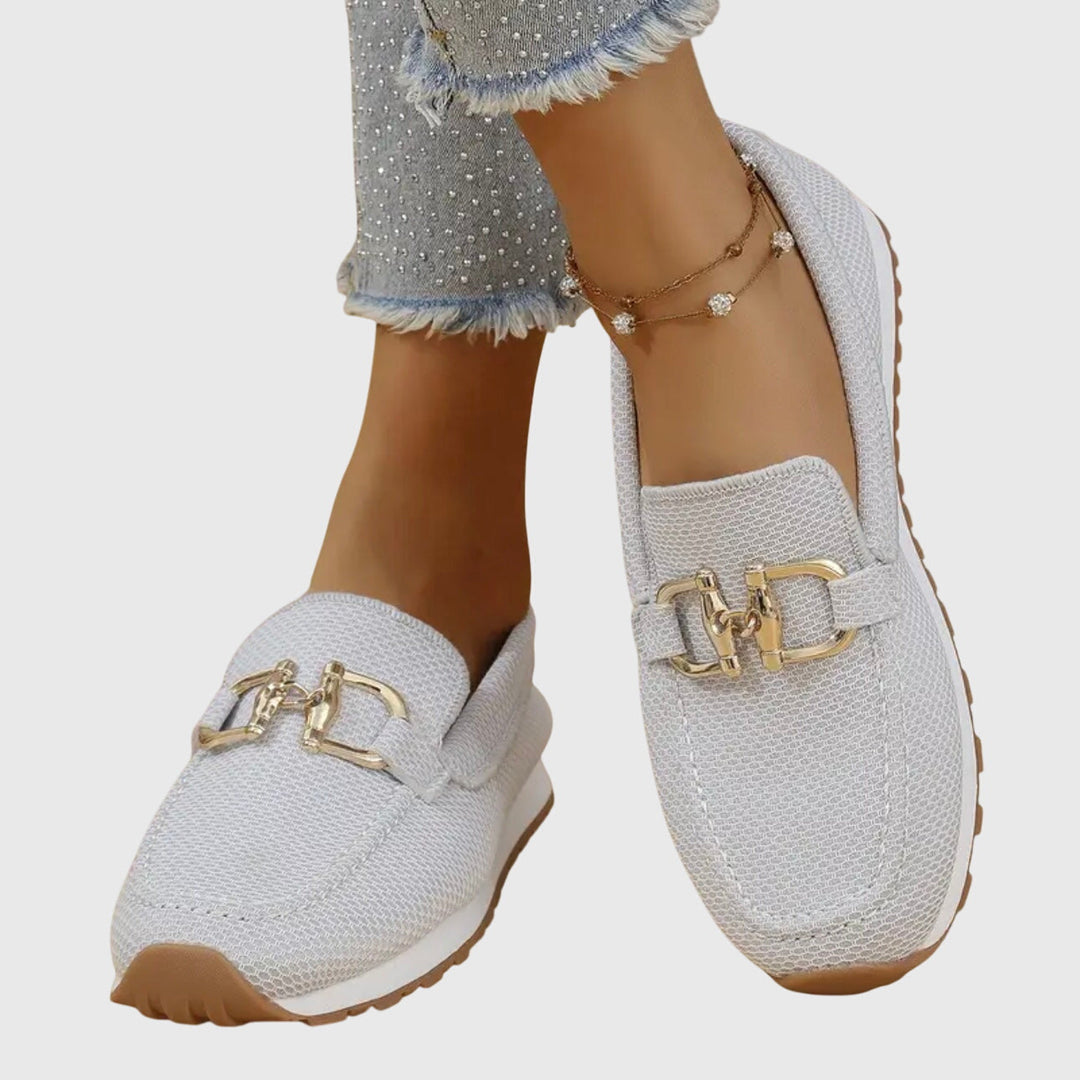 White loafers with gold buckles worn by a person on a light gray background