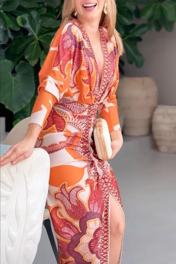 Woman wearing an orange and pink patterned dress sitting on a white chair with green plants in the background.