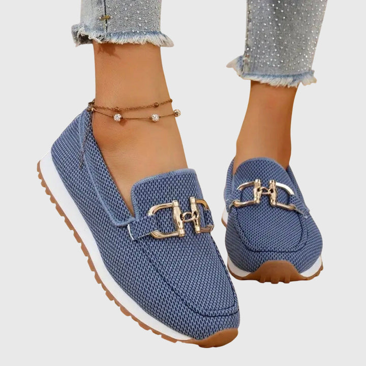 Blue loafers with gold buckles worn by a person on a light gray background