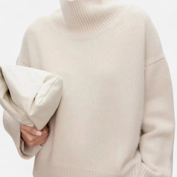 Person wearing a beige turtleneck sweater holding a matching pillow against a white background