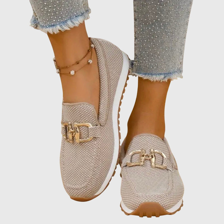 Beige loafers with gold buckles worn with light blue jeans on a white background
