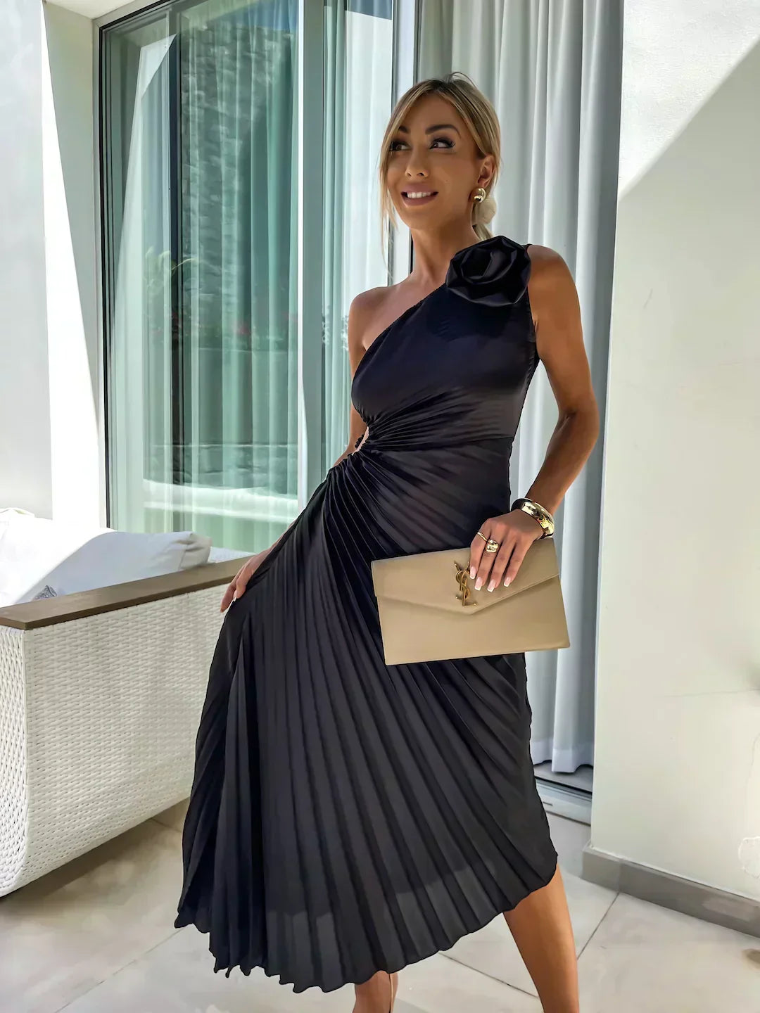 Woman in a black pleated dress holding a beige clutch in an indoor setting.