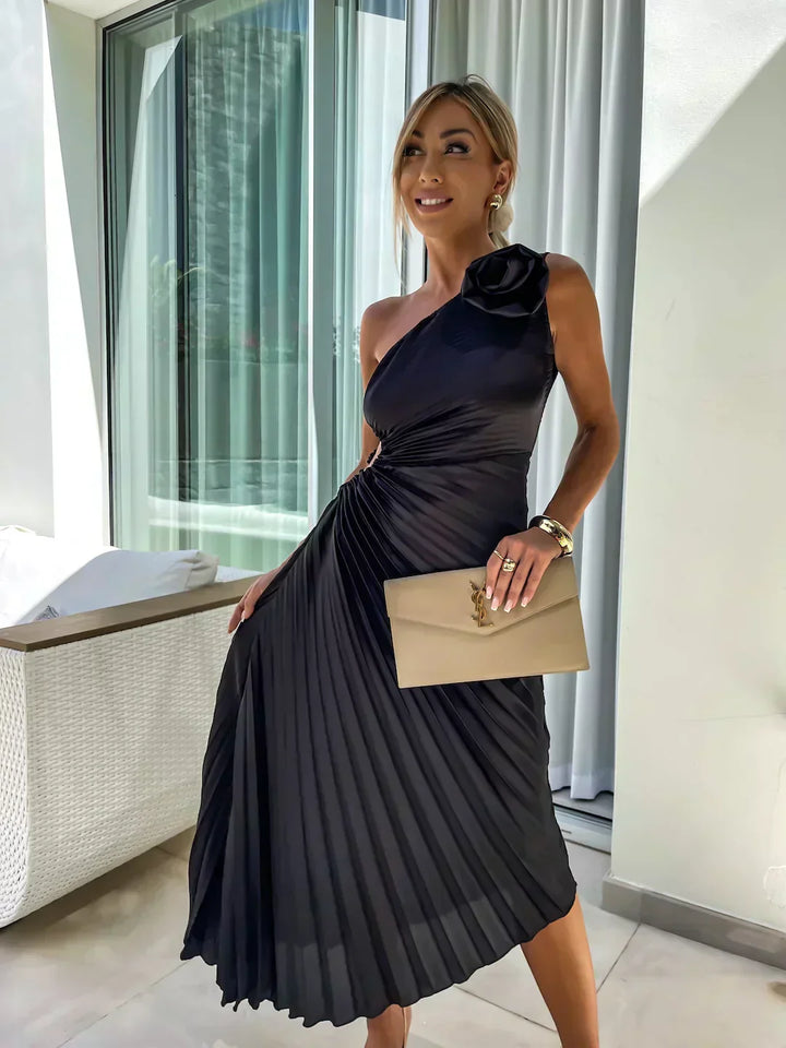 Woman in a black pleated dress holding a beige clutch in an indoor setting.