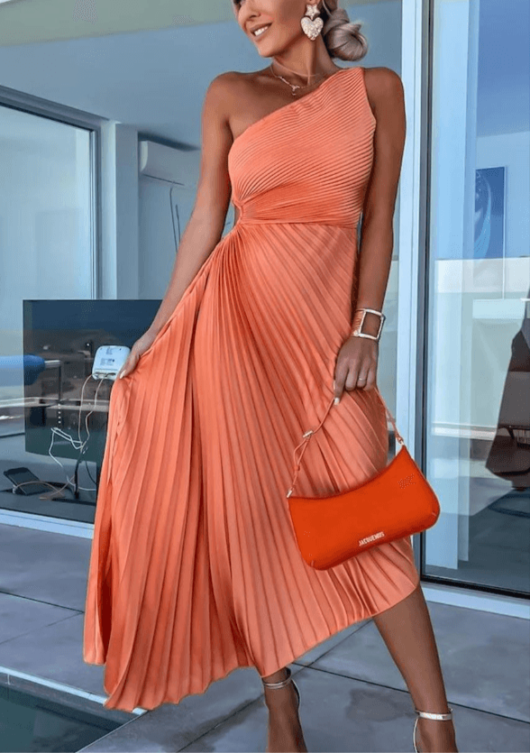 Woman in an orange pleated dress holding an orange clutch in a modern indoor setting.
