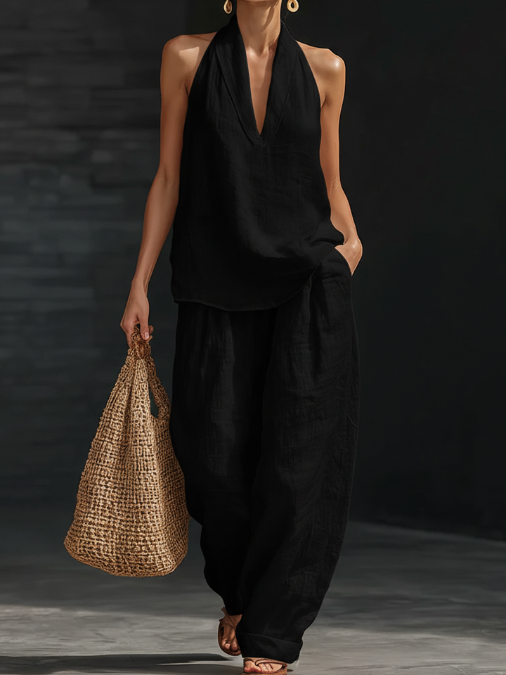 Woman in black halter top and wide-leg pants holding a woven bag against a dark background