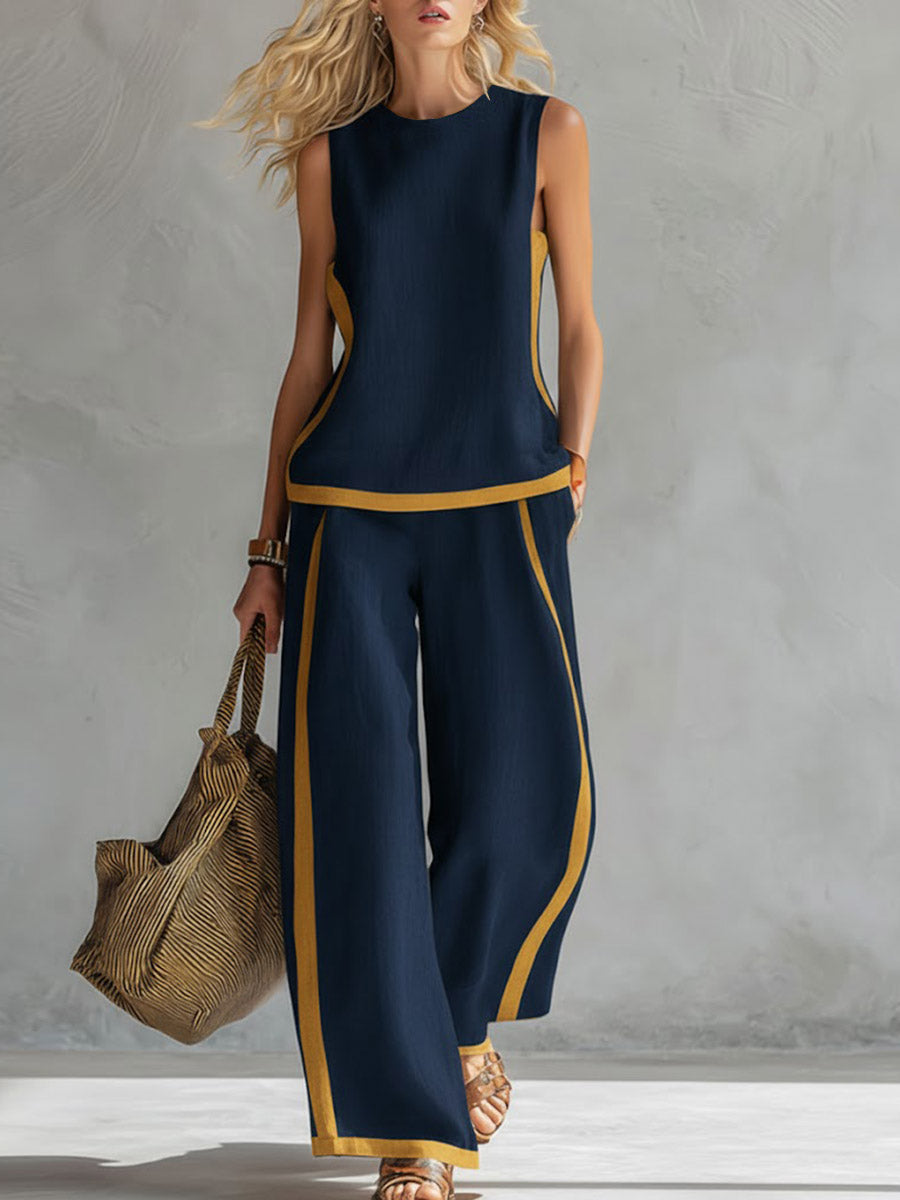 Woman wearing a navy sleeveless top and wide-leg pants with yellow accents, holding a woven bag against a gray background.