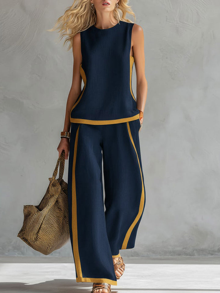 Woman wearing a navy sleeveless top and wide-leg pants with yellow accents, holding a woven bag against a gray background.