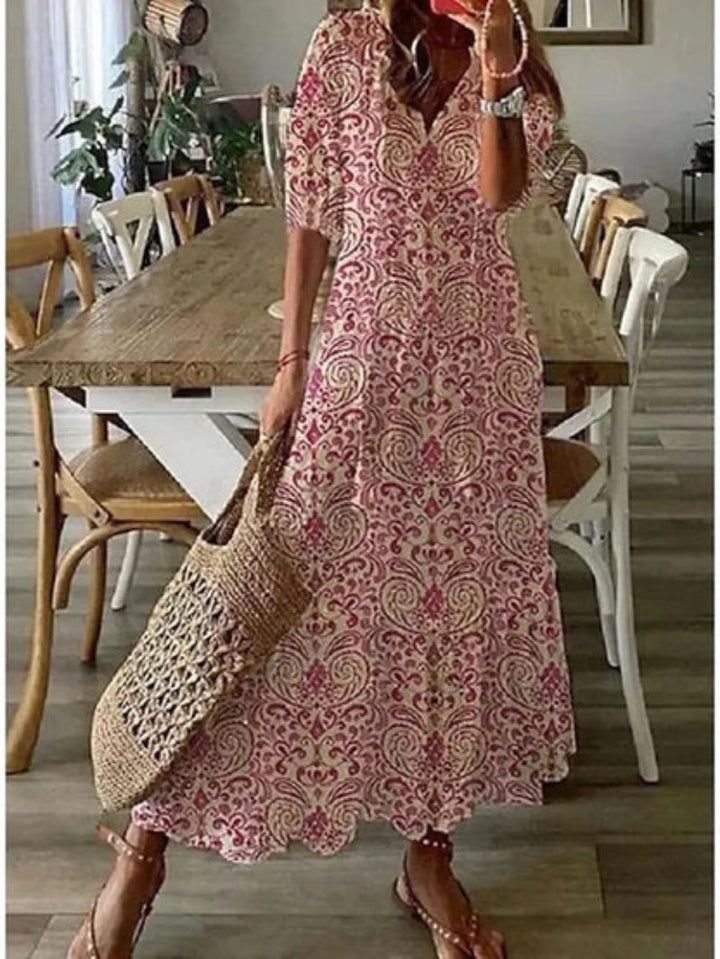 Woman in a pink patterned dress standing in a dining room with wooden table and chairs.