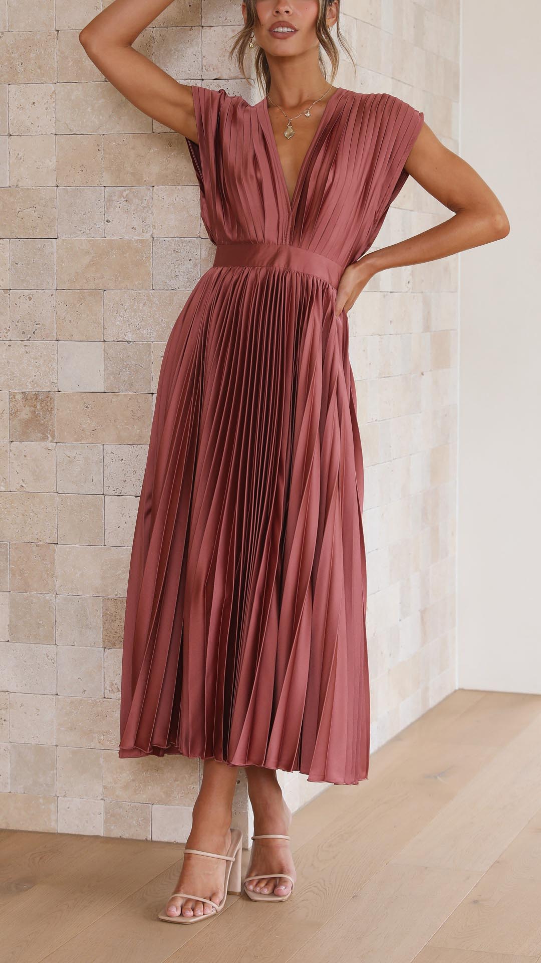 Woman wearing a rust-colored pleated dress against a beige tiled wall.