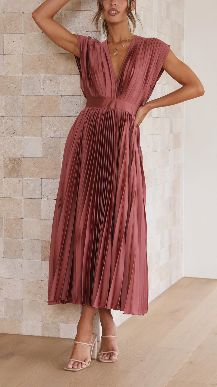 Woman wearing a rust-colored pleated dress against a beige tiled wall.